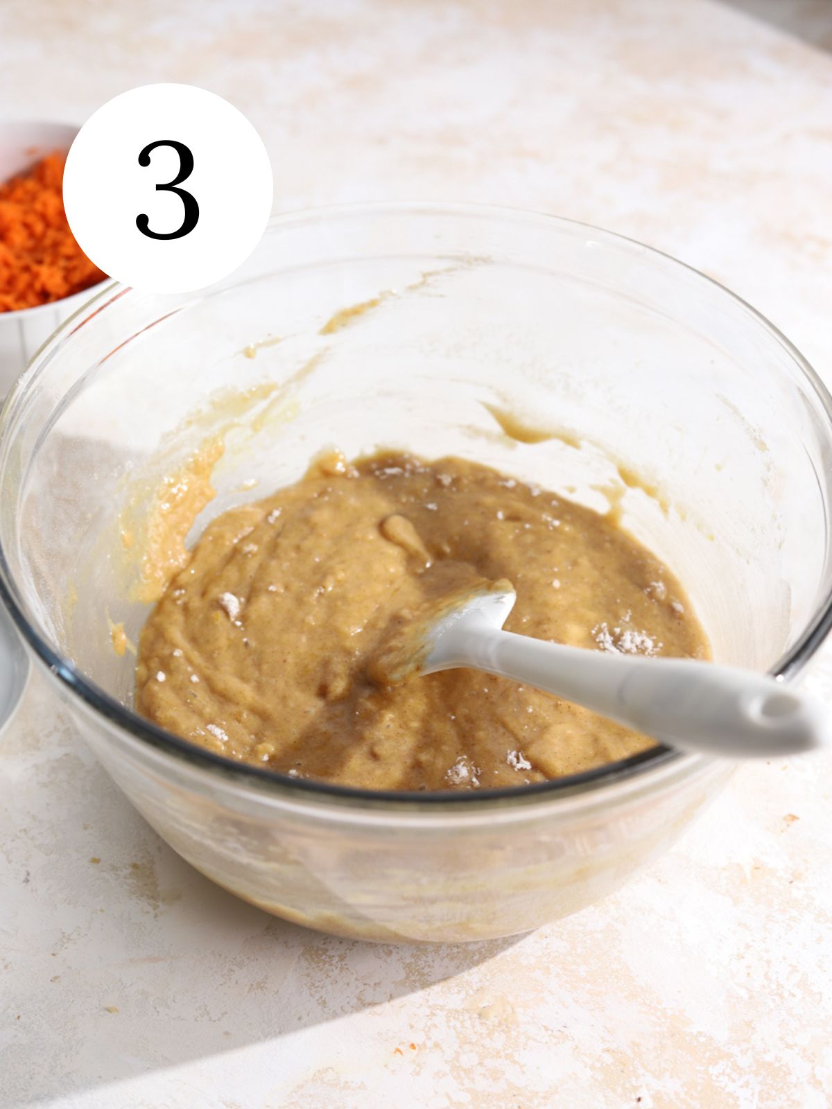 A glass bowl with flour stirred into batter with a spatula.