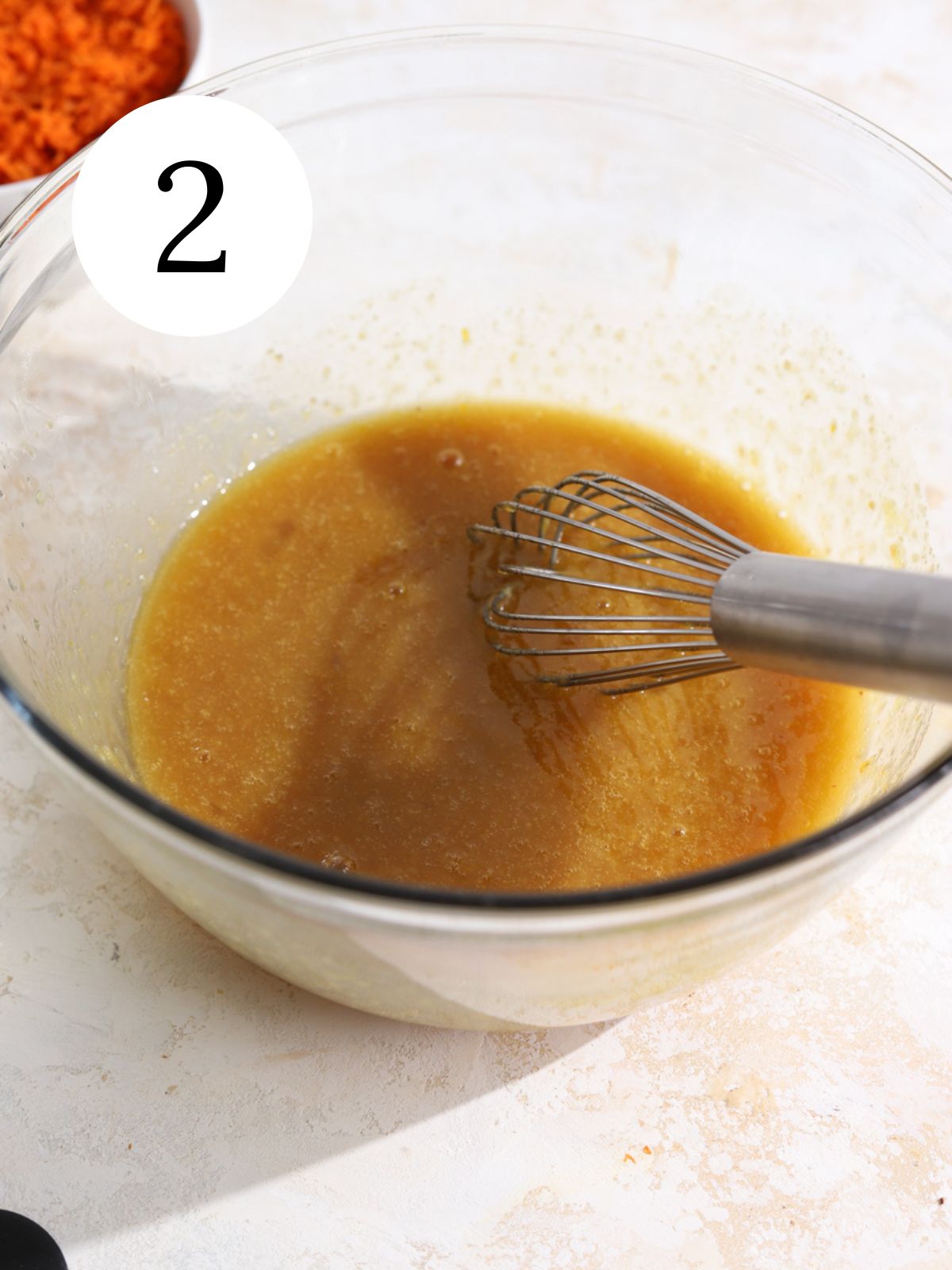 Wet ingredients in a bowl with a wisk and a bowl of shredded carrots.