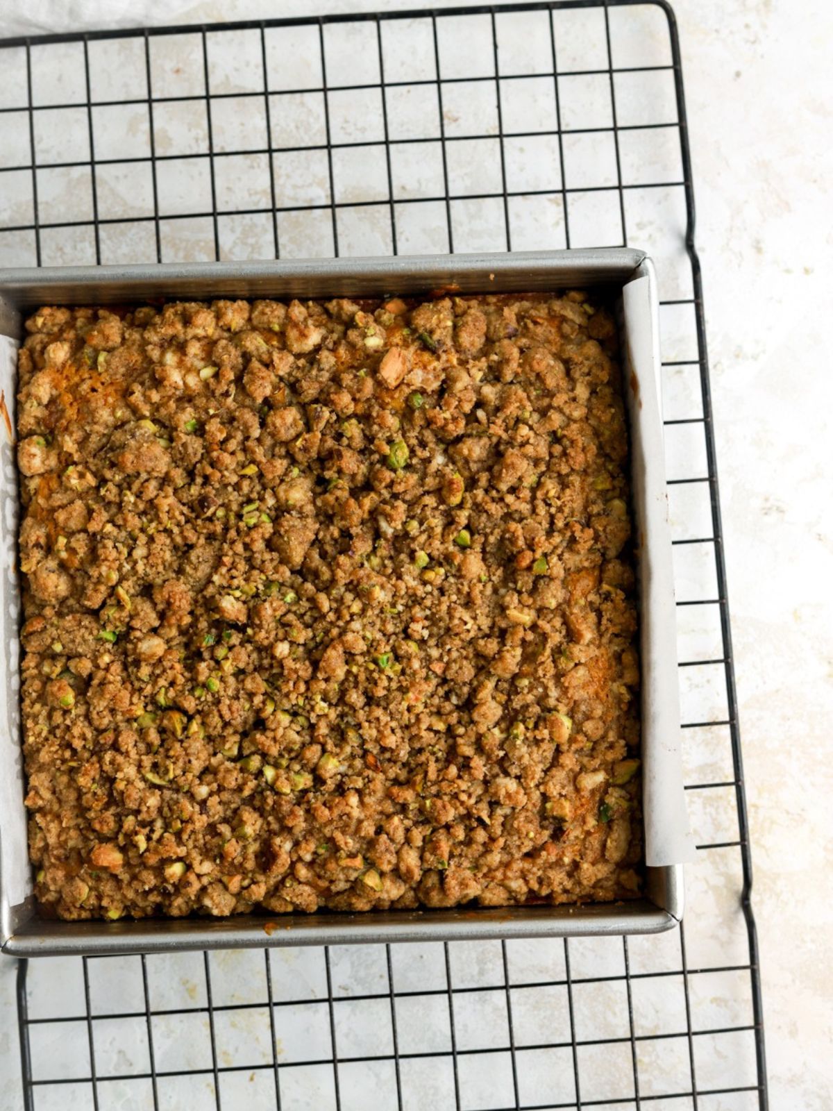A square pan with a cake with pistachio streusel on a wire rack. 