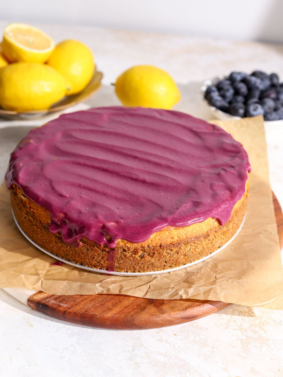 Cheesecake topped with purple blueberry curd, a bowl of lemons and bowl of blueberries.