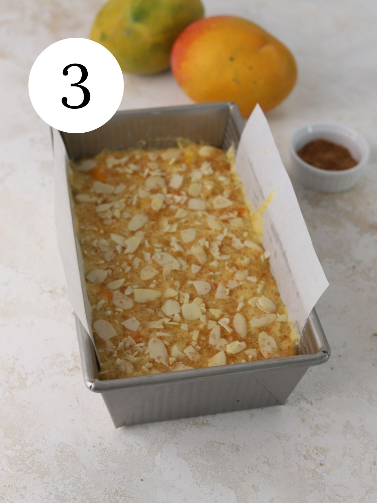 Cake batter in a loaf pan next to two mangoes.