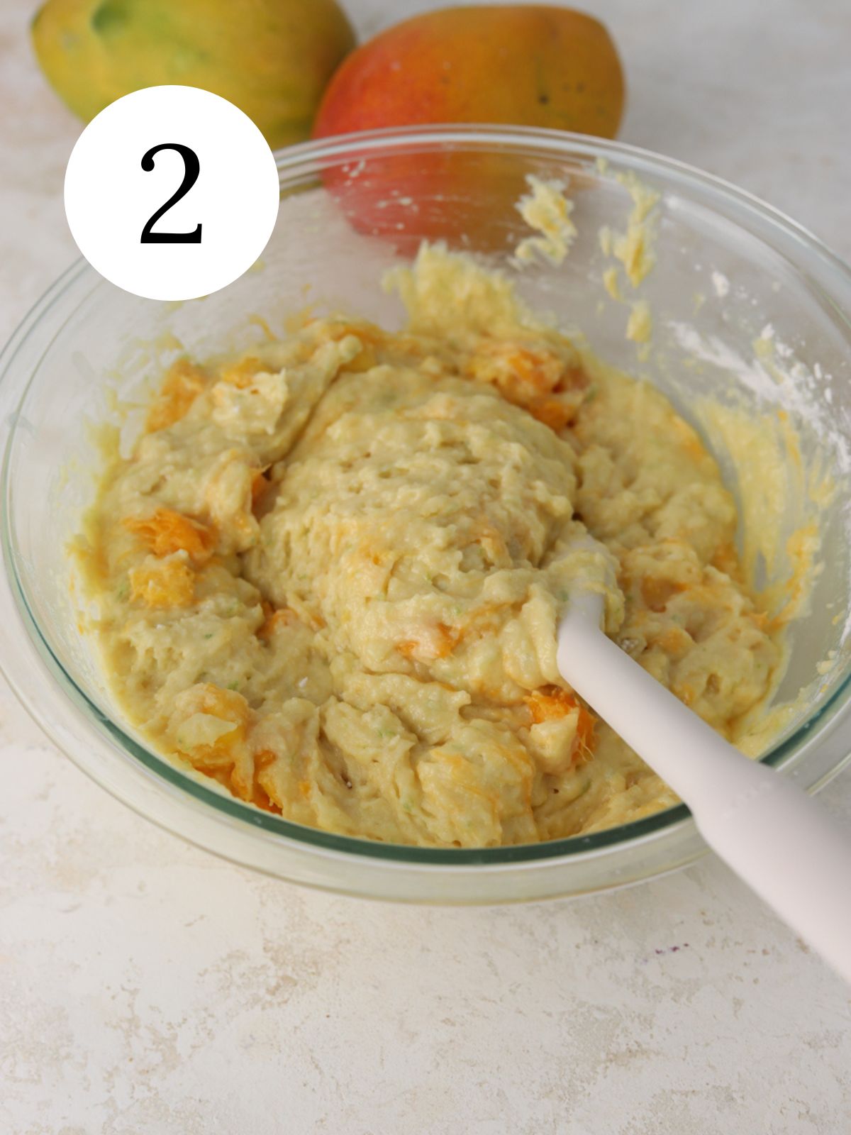 Mango cake batter in a bowl with a spatula next to two mangos.