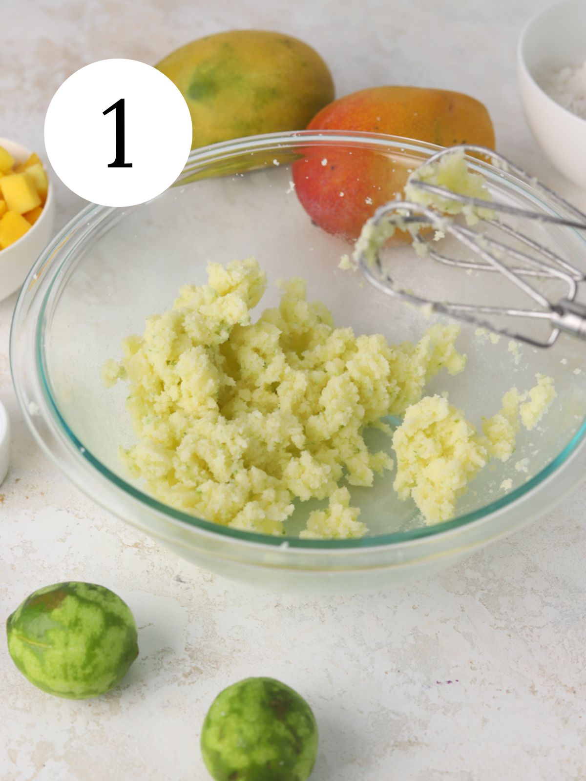 Butter and sugar mixed together in a bowl with an electric mixer and some limes and mangoes.