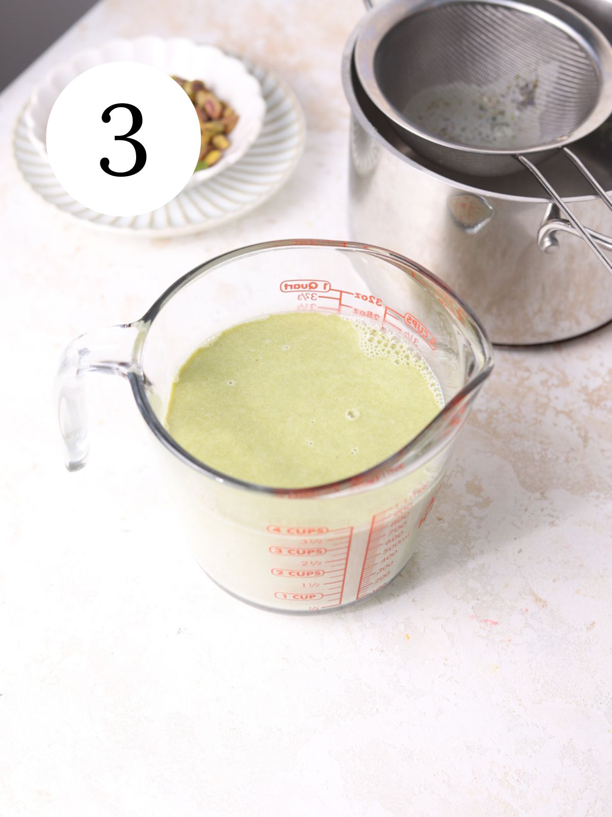 Pistachio panna cotta in a glass mixing bowl next to a pot with a sieve.