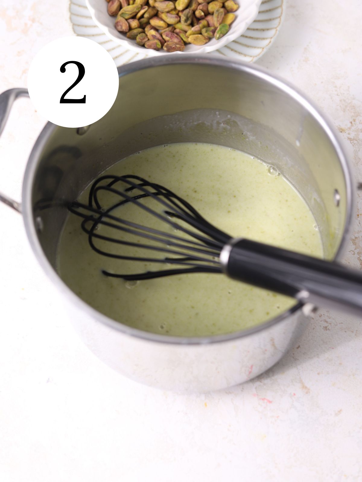 Pistachio paste mixed into cream in a pot with a whisk and a bowl of pistachios.