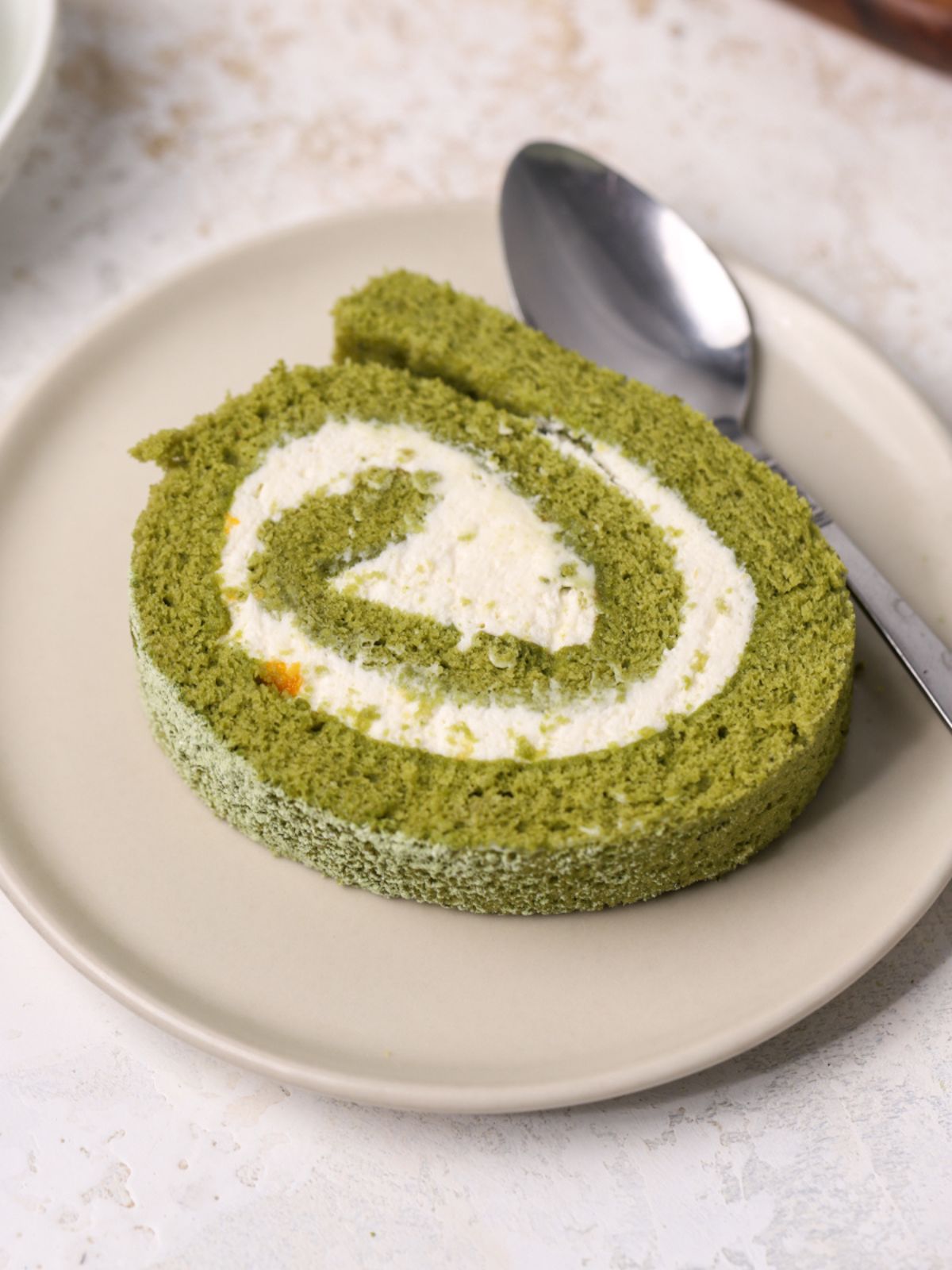 A slice of matcha roll cake with cream filling on a plate with a spoon.