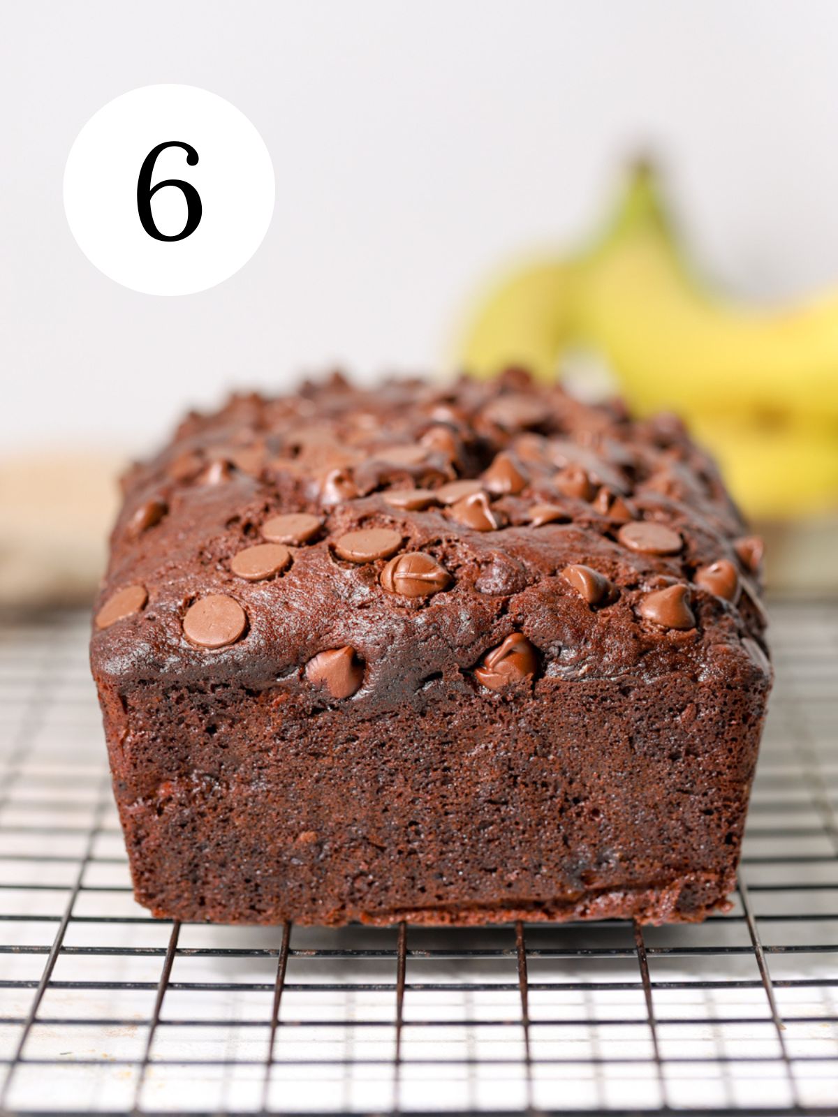 A chocolate chip banana bread on a rack in front of bananas. 