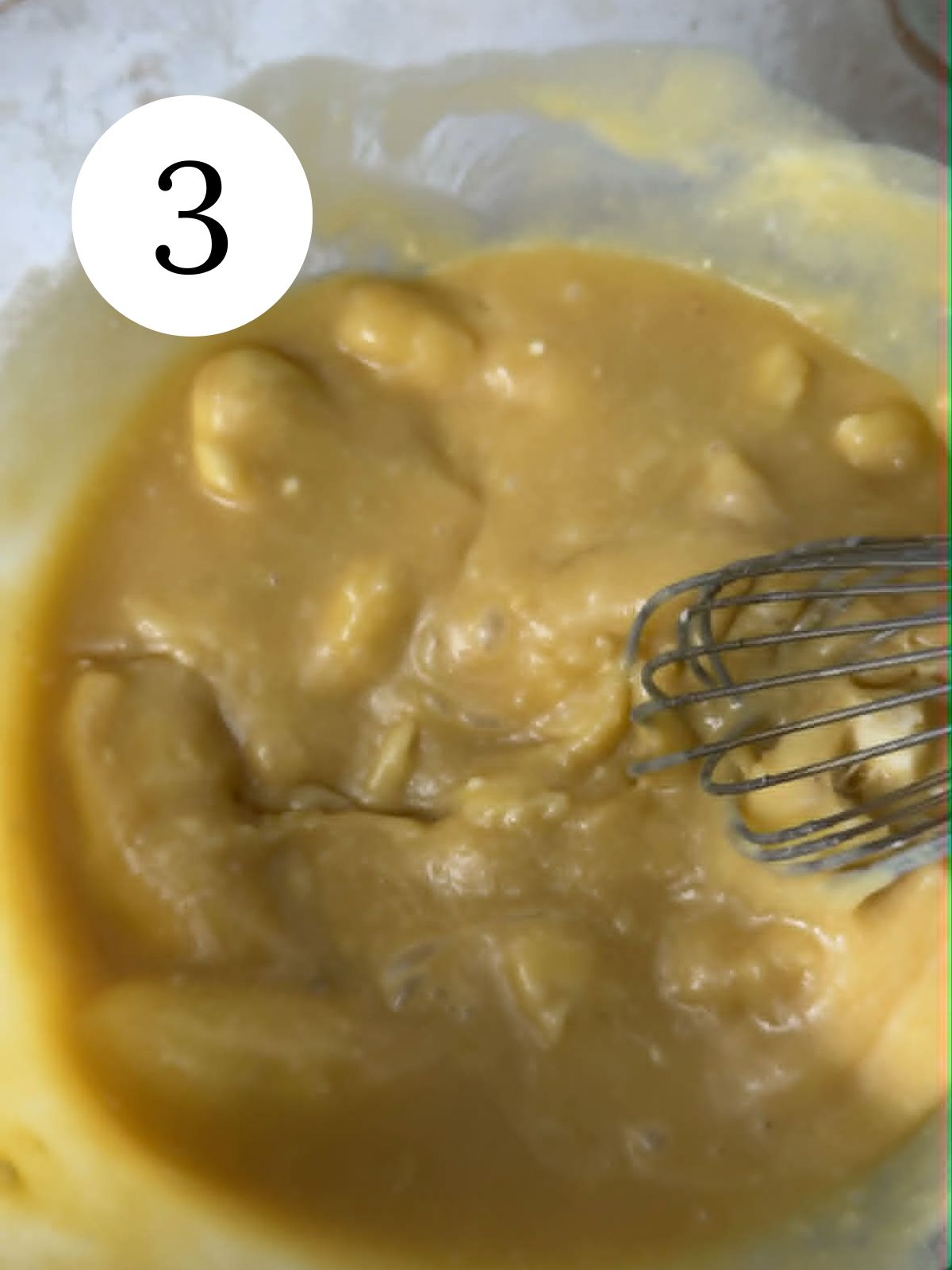 A whisk mixing bananas into batter in a bowl.