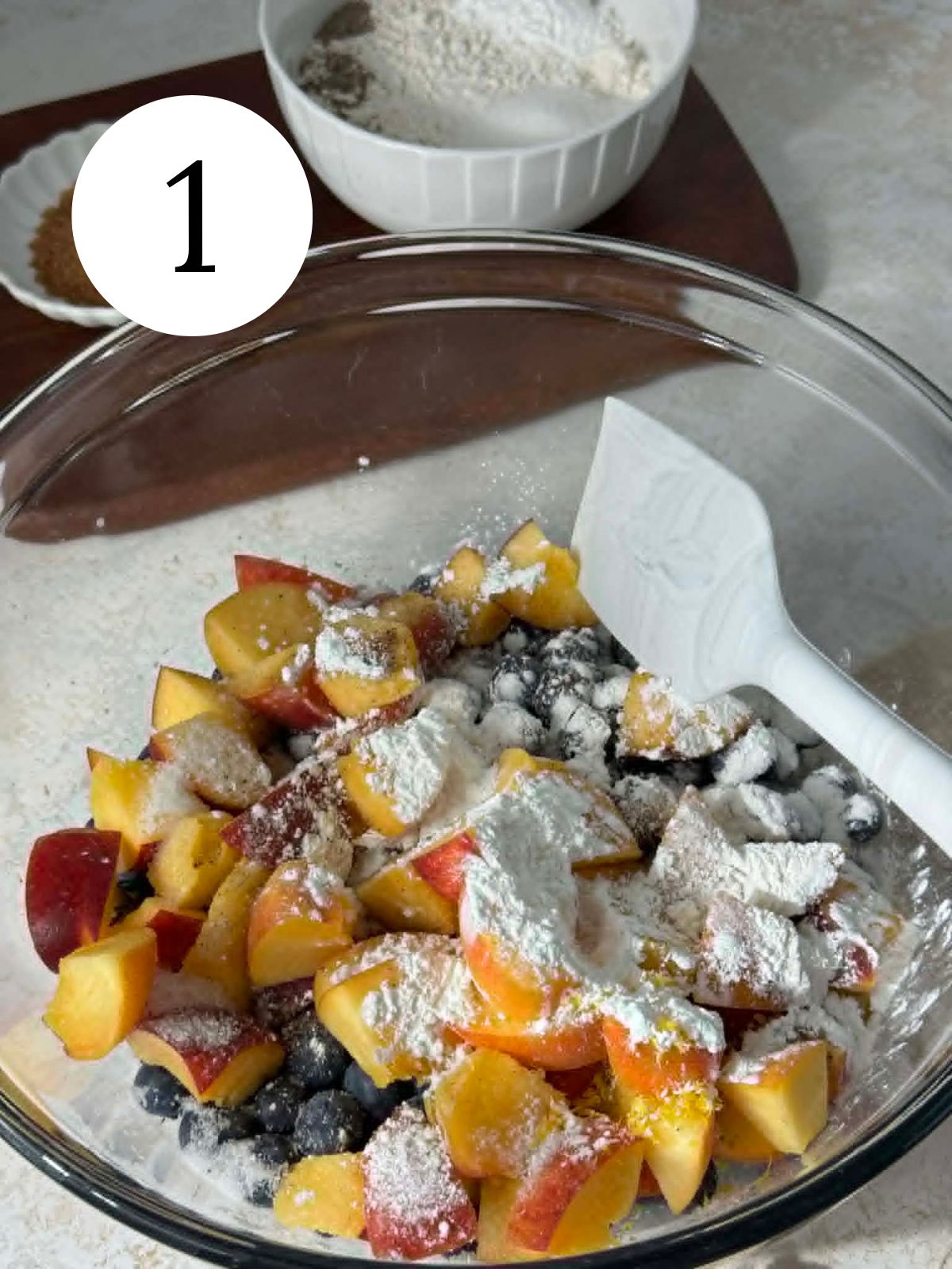 A glass mixing bowl with cornstarch sprinkled over peaches and blueberries.
