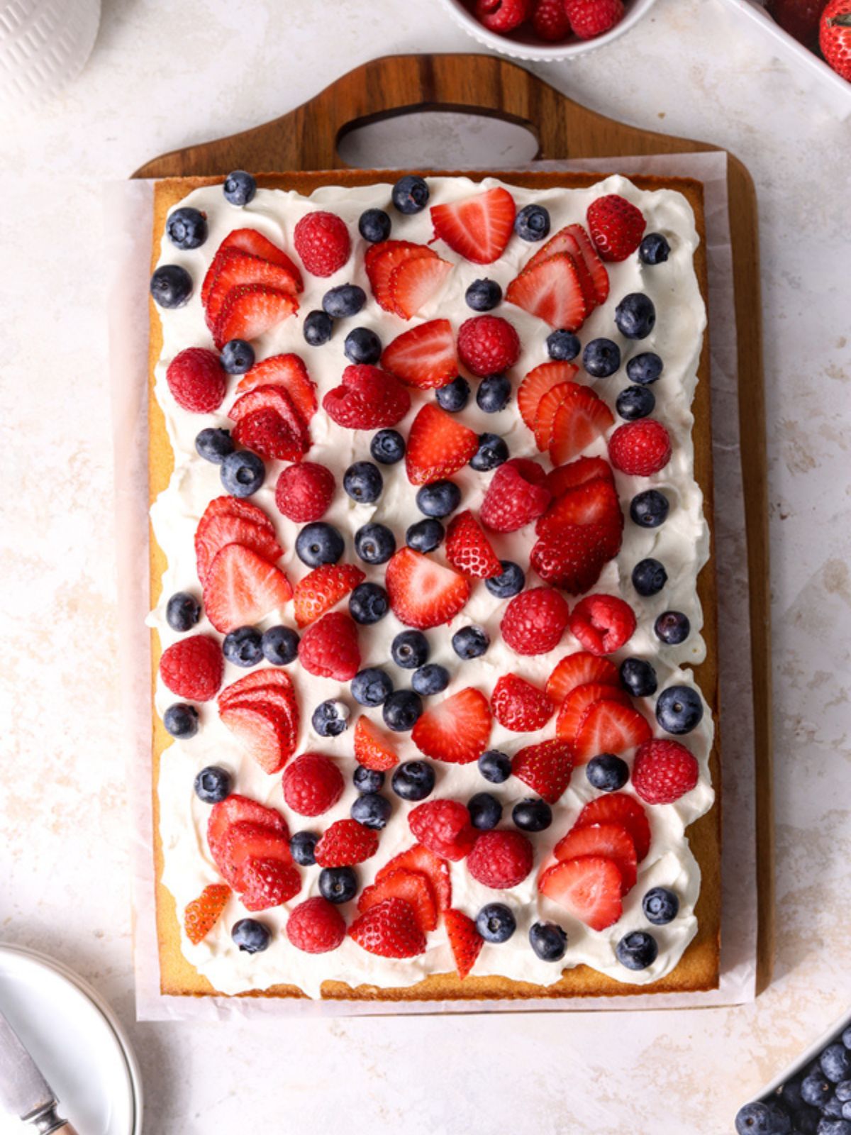 A sheet cake with whipped cream and fresh berries on a wooden tray.